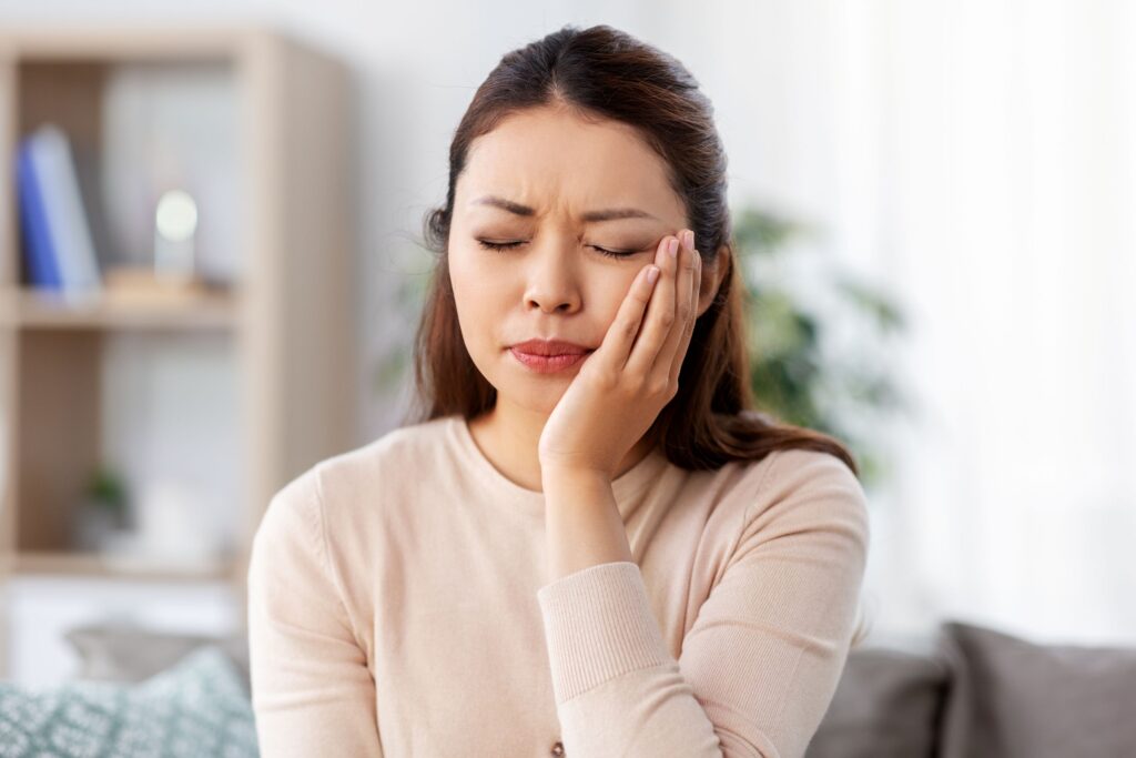 Woman in beige sweater holding hand to cheek in pain